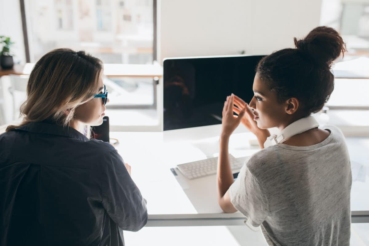 Indoor portrait from back of two girls sitting in front of computer with black screen. African female office worker in headphones talking with colleague beside window.. Digital speech therapy